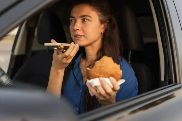 Close-up of woman using mobile phone, eating burger and talking on the speaker while driving car. Beautiful woman talking on in-car speakerphone. Copy space