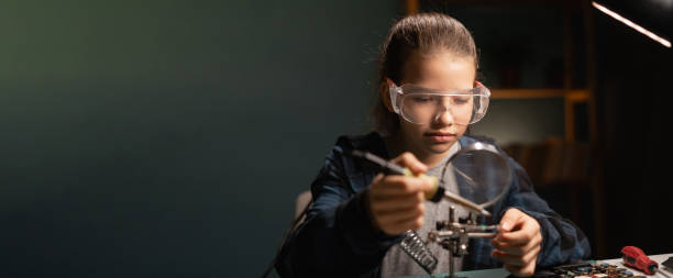 istock Cute girl in safety glasses, learning how to solder microprocessor cables and printed circuits uses a magnifying glass, repairing electronics 1782935013