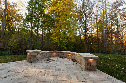 Picturesque backyard view in autumn season with patio pavers and stone wall, autumn leaves, and colorful woods in the background.