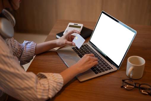 Cropped side view image of a woman using her laptop and holding her credit card, registering her credit card account on a shopping website or online banking website. A white-screen laptop mockup