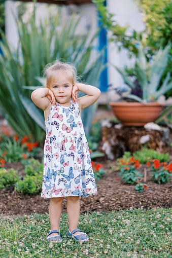 Little girl stands thoughtfully in the garden by the flower bed, touching her face with her hands. High quality photo