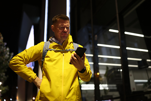 Male tourist with backpack looks for taxi number on phone standing on street illuminated with light. Focused man holds gadget in hand looking at screen