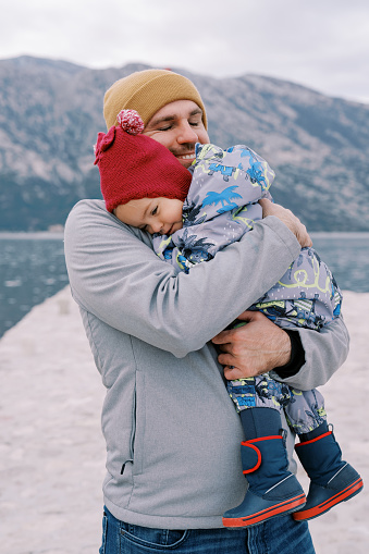 Smiling dad cuddles a little girl on a pier against the backdrop of mountains. High quality photo