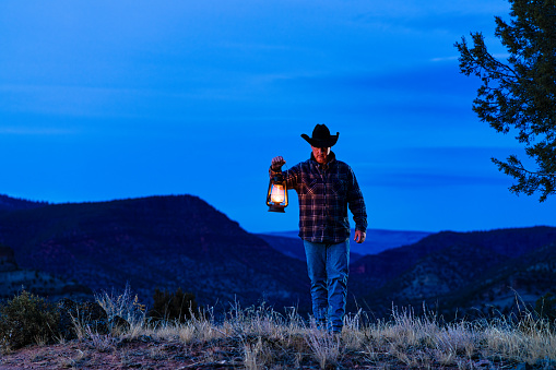 Cowboy Rancher Carrying Old Style Lantern - Man in natural outdoor Western USA area with mountain backdrop captured at dusk during blue hour.