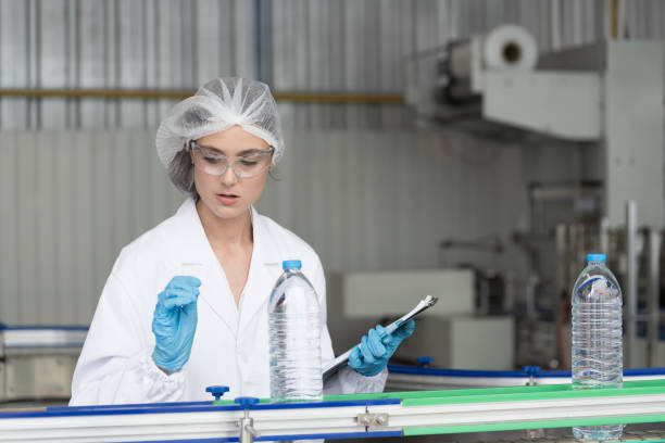 Female worker wearing uniform and hairnet inspecting quality of purified drinking plastic bottled water inside automated conveyor belt production line in drinking water factory trabajadora vestida con uniforme y redecilla para el cabello inspeccionando la calidad del agua embotellada de plástico potable purificada dentro de la línea de producción automatizada de cintas transportadoras en la fábrica de agua potable
