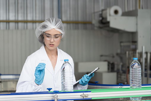 Female worker wearing uniform and hairnet inspecting quality of purified drinking plastic bottled water inside automated conveyor belt production line in drinking water factory