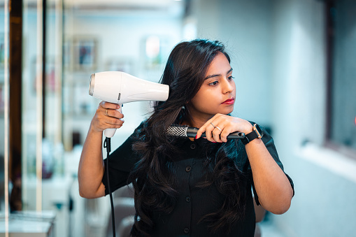 Portrait of Female hairdresser at the salon holding styling her hair using hair dryer and Round Hair Brush. Woman hairdresser in happy mood styling her hair at salon,
