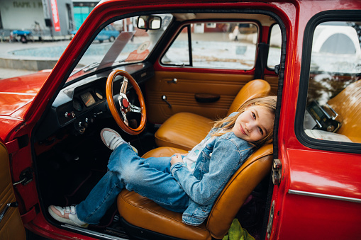 Teenage girl in denim clothes driving a red vintage retro car. Exhibition of a collection of old cars. Walks with children.