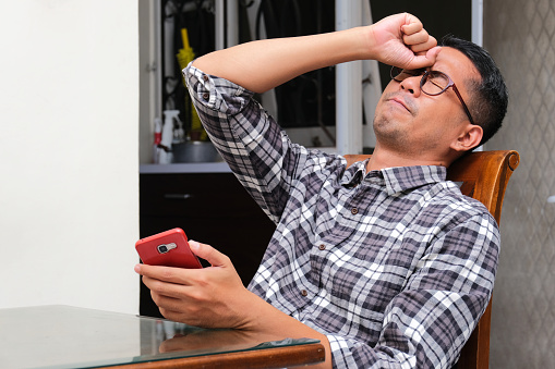 Adult Asian man sitting and showing stress expression while holding mobile phone