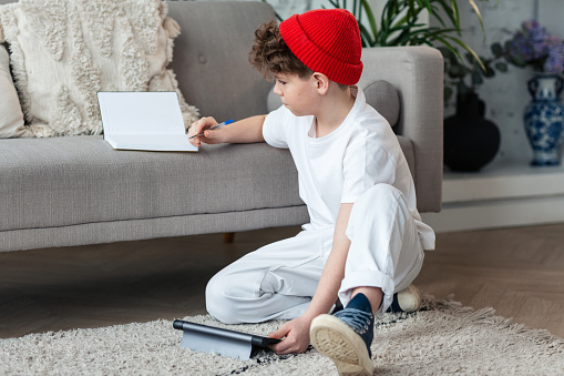 A teenager boy in a red cap uses a digital tablet while sitting on the floor at home for the purpose of learning, communicating in social networks, searching for information, online shopping.