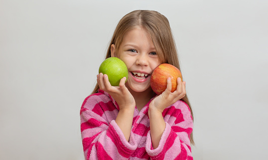 Portrait of caucasian little girl with open wide smile in bathrobe holding green and red apples in hands looking at camera on white background