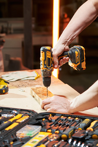 Worker's hands on a workbench with different tools drill a wooden part using a cordless drilldriver