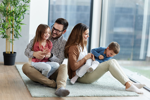 Cheerful parents having fun while tickling their small kids on carpet in the living room.