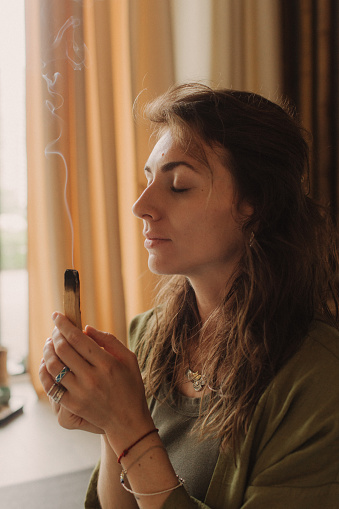 A young woman holds a smoking Palo Santo stick in her hands. Buddhist healing practices. Clearing the space of negative energy. Aromatherapy. Selective focus,close up. Soft focus and noise effect