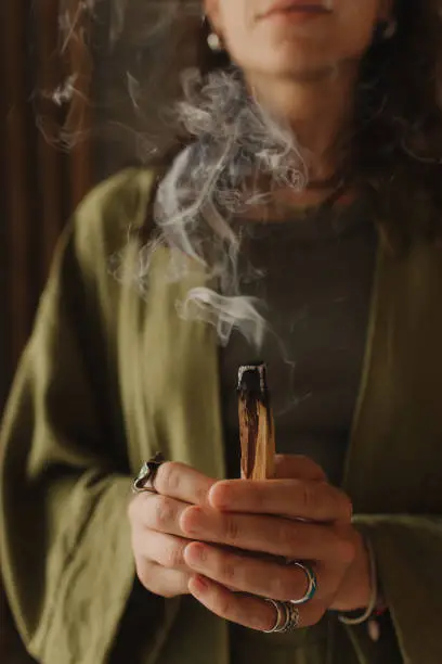 A young woman holds a smoking Palo Santo stick in her hands. Buddhist healing practices. Clearing the space of negative energy. Aromatherapy. Selective focus,close up. Soft focus and noise effect