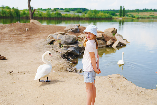 Cute girl in blue panama hat standing on the shore of the lake with swans.