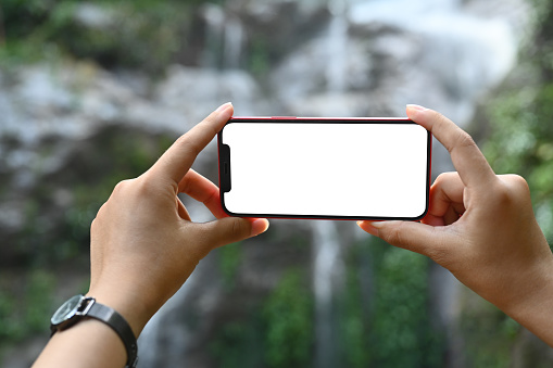 Woman hand holding smart phone in hand on the blur background of waterfall.