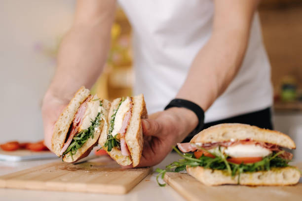 Handsome young man slice Italian sandwich and show what inside. Delicious lunch at home stock photo