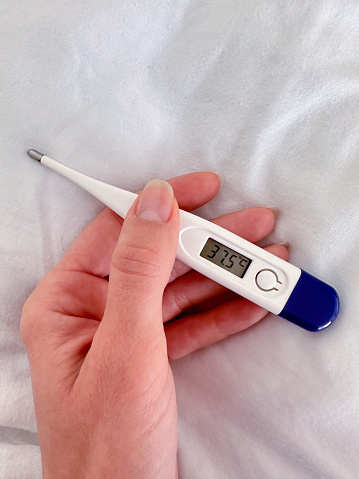 An electronic thermometer with a temperature of 37.5 in a female hand on a white blanket background