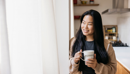 Young beautiful woman with a mug drinking coffee next to a window in a kitchen. Smiling woman enjoying a coffee break at home.