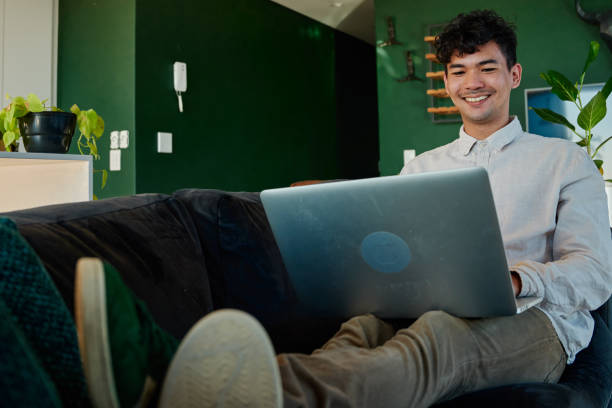 istock Young multiracial man in long sleeved shirt smiling while using laptop on sofa at home 1783534856