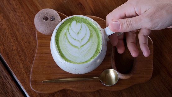Top down view of a beautifully served cup of matte green tea with an ingenious pattern on the milk froth. The cup stands on a wooden tray in the same color scheme as the table.