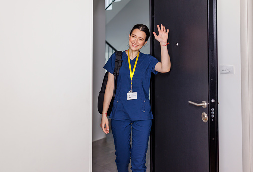 Young Caucasian female nurse coming home from work with a smile on her face. She is standing in her scrubs, while carrying her backpack on her shoulder and waving hello.