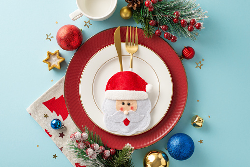 Top view of playful Christmas party table arrangement featuring amusing cutlery pocket, elegant plates, napkin, cozy mug, festive baubles, frost-kissed fir twigs, holly berries, pastel blue backdrop