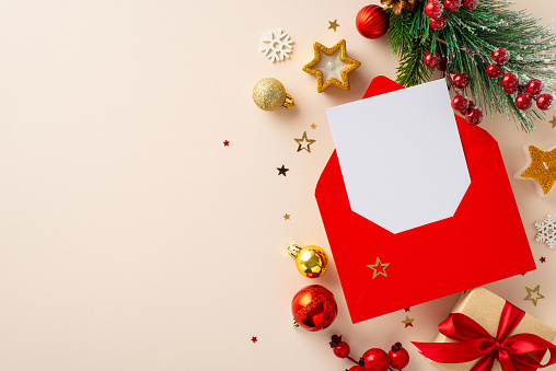Get ready for holidays with wish list in red envelope surrounded by festive elements like gift box, gold ornaments, fir branch, holly berries, snowflakes on pastel beige backdrop with space for advert