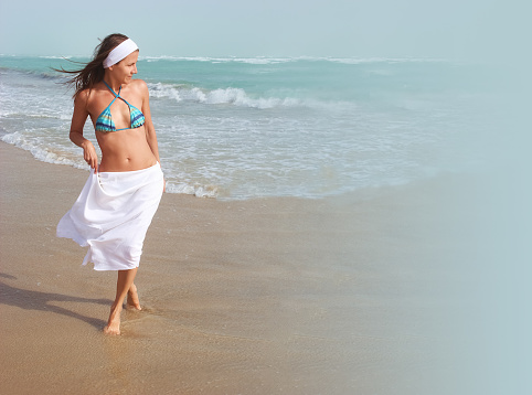 Beautiful girl walks barefoot, on sandy beach, along ocean, wind is blowing her hair and white skirt