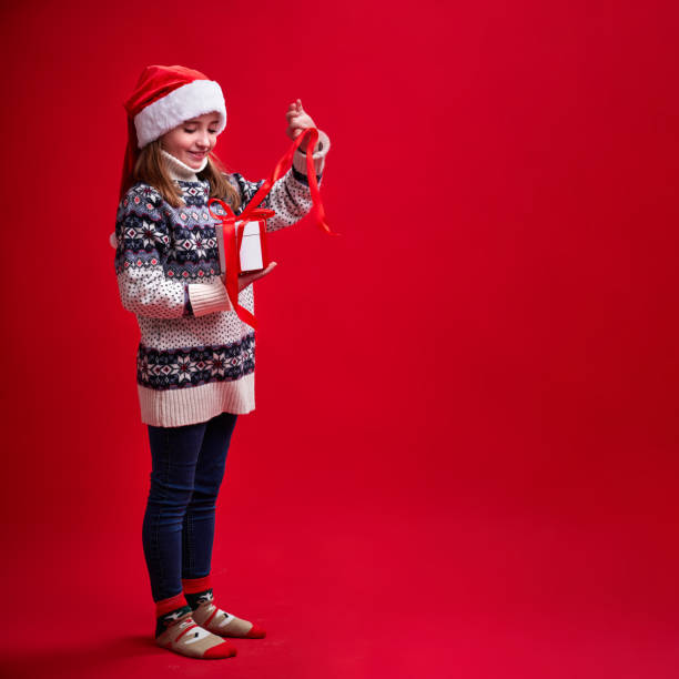 Happy cheerful girl in sweater and Santa's hat opens gift on red background stock photo