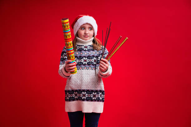 Funny girl in Santa's hat holds sparklers and cracker on red background. stock photo