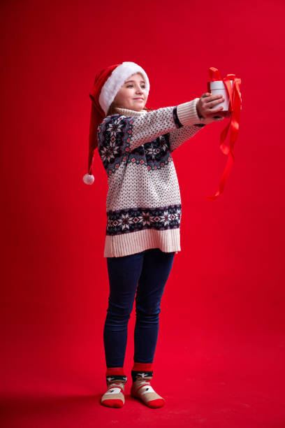 Happy cheerful girl in sweater and Santa's hat holds gift on red background stock photo