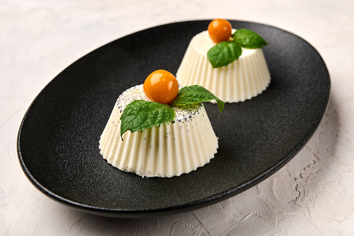 Two panna cotta with natural vanilla pod, physalis or winter cherry and mint on black oval plate