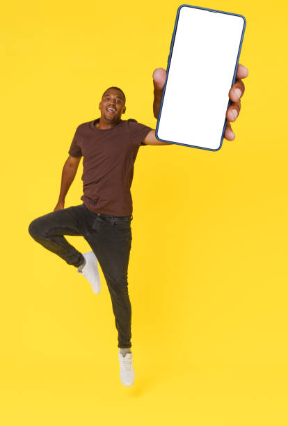 istock Energetic African American student is captured jumping with mobile phone featuring empty white screen in hand, on yellow background. This image is perfect for product placement or advertising concepts. 1783602886