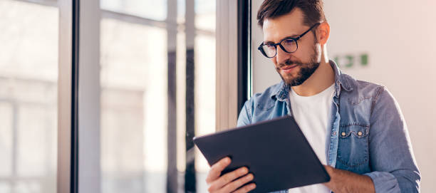 istock Smiling young businessman working online with digital tablet while standing by window. 1783604167