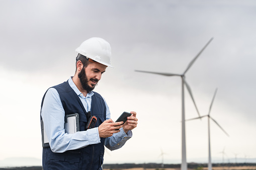 Professional male engineer wearing safety helmet, using smartphone at outdoors wind turbine park. Engineering and safety at work