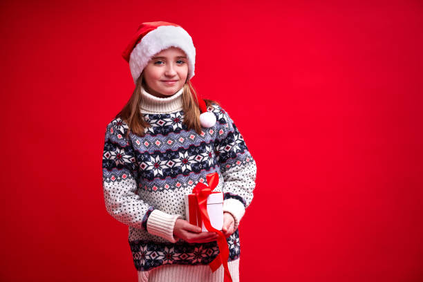 Happy cheerful girl in sweater and Santa's hat holds gift on red background stock photo