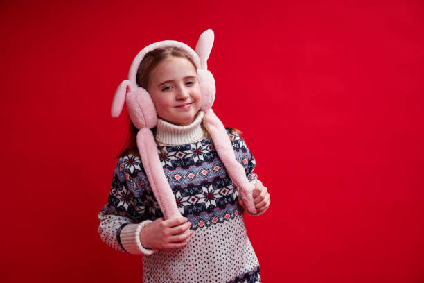 Cute girl in sweater and fluffy headphones smiles on red background stock photo