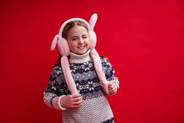 Girl in sweater and fluffy headphones smiles and winks on red background stock photo