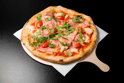 Tasty pizza with prosciutto di parma, arugula, halved cherry tomatoes, chicken meat, pineapple and dried herbs served on wooden board . Italian traditional dish. Delicious food