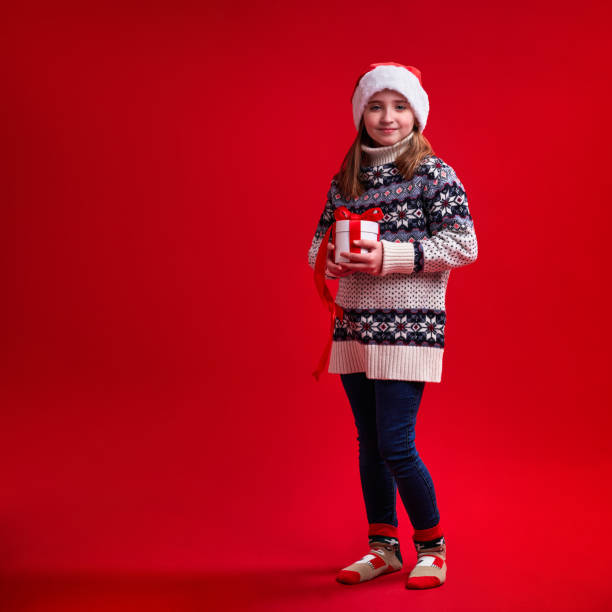 Happy cheerful girl in sweater and Santa's hat holds gift on red background stock photo