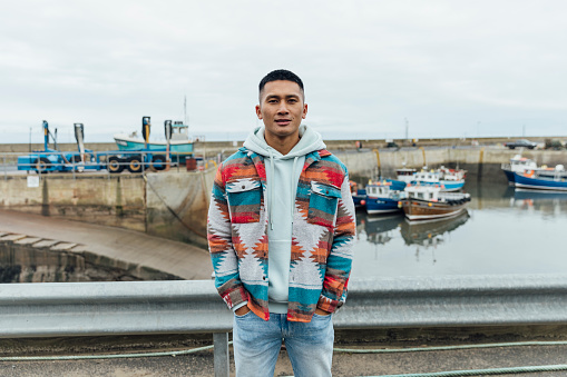 A portrait shot of a man in the seaside town of Seahouses in Northumberland. They are wearing warm, casual clothing on an overcast winter day. They stand, smile and look at the camera.