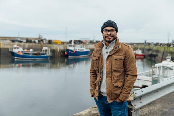 A portrait shot of a man in the seaside town of Seahouses in Northumberland. They are wearing warm, casual clothing on an overcast winter day. They stand, smile and look at the camera.