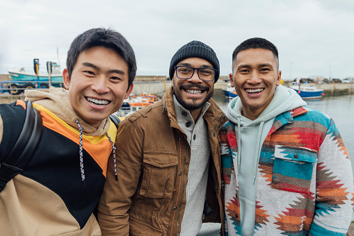 A selfie shot of a multiracial group of three male friends in the seaside town of Seahouses in Northumberland. They are wearing warm, casual clothing on an overcast winter day. They look and smile at the camera.