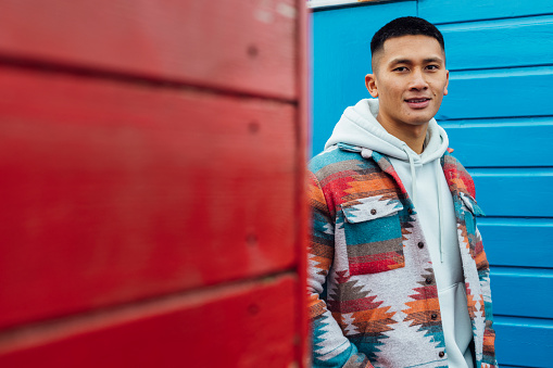 A portrait shot of a man in the seaside town of Seahouses in Northumberland. They are wearing warm, casual clothing on an overcast winter day. They stand in front of a beach hut as they smile and look at the camera.