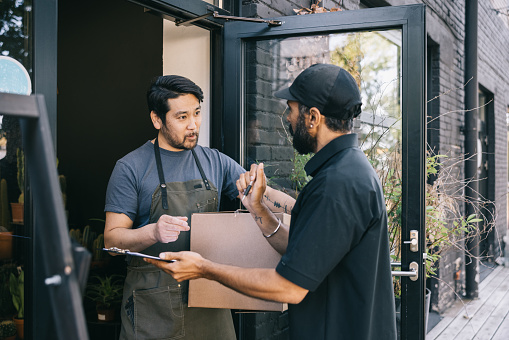 A small business owner stands at the entrance of their shop, receiving a package from a courier, an essential moment in the day-to-day operations of their business.