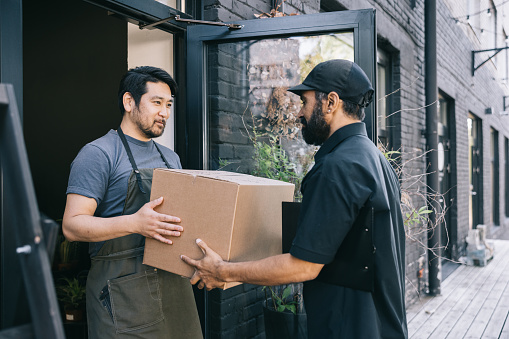 A small business owner stands at the entrance of their shop, receiving a package from a courier, an essential moment in the day-to-day operations of their business.