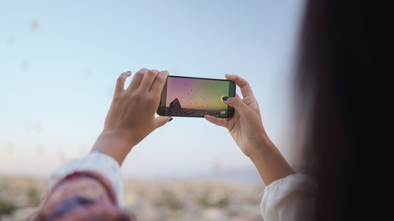 A young female tourist is taking photos of hot air ballons with her mobile smart phone from the balcony of the hotel during her travel.