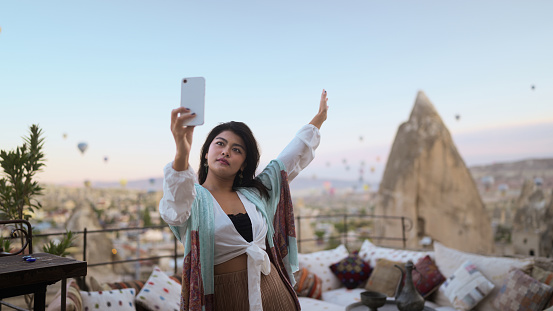 A young female tourist is taking selfies wither her mobile smart phone in the balcony of a cave hotel in Cappadocia during her travel.
